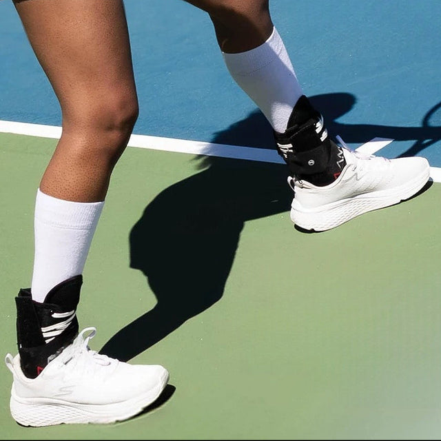 Person wearing white sneakers and the AAMO ankle brace on a tennis court.